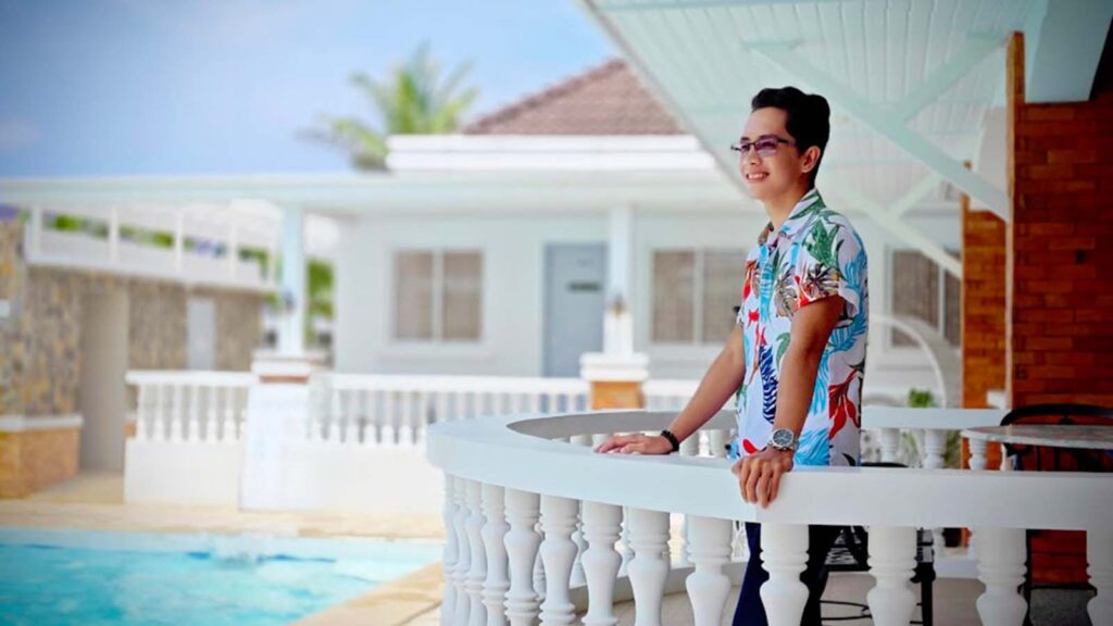 Alpha Jun Ibieza standing on a white balcony overlooking a pool, wearing a tropical floral shirt, glasses, and a watch. Smiling and looking into the distance in a bright sunny resort setting in the Philippines. This is the face behind alphajun.com.
