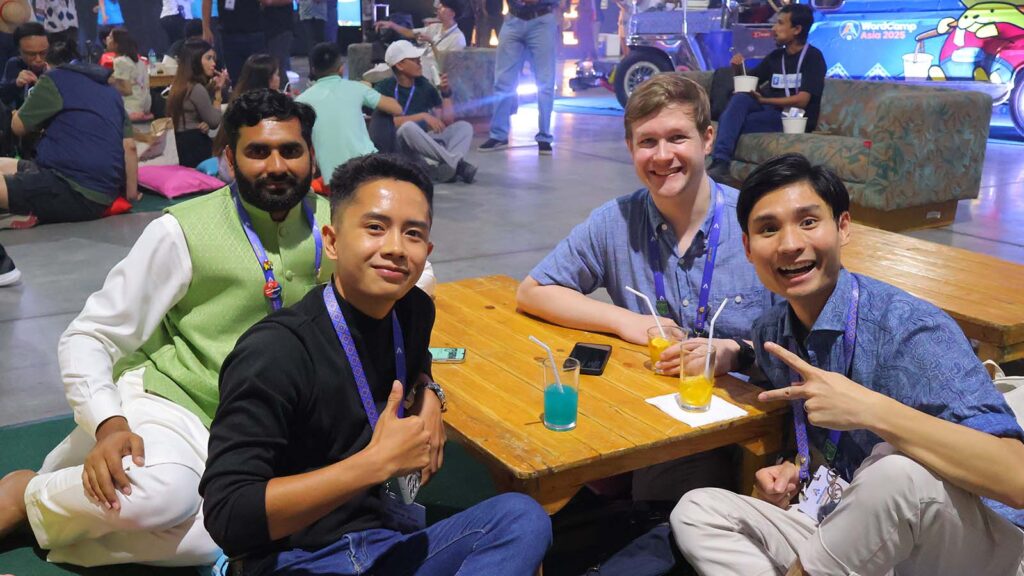 People networking and relaxing at a WordCamp event lounge area, sitting around a table with drinks and chatting with other attendees.