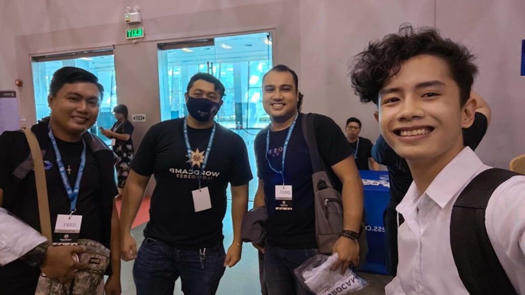 Group of WordPress community attendees sitting around a wooden table with drinks, wearing event lanyards and smiling during a social gathering at a WordCamp event.